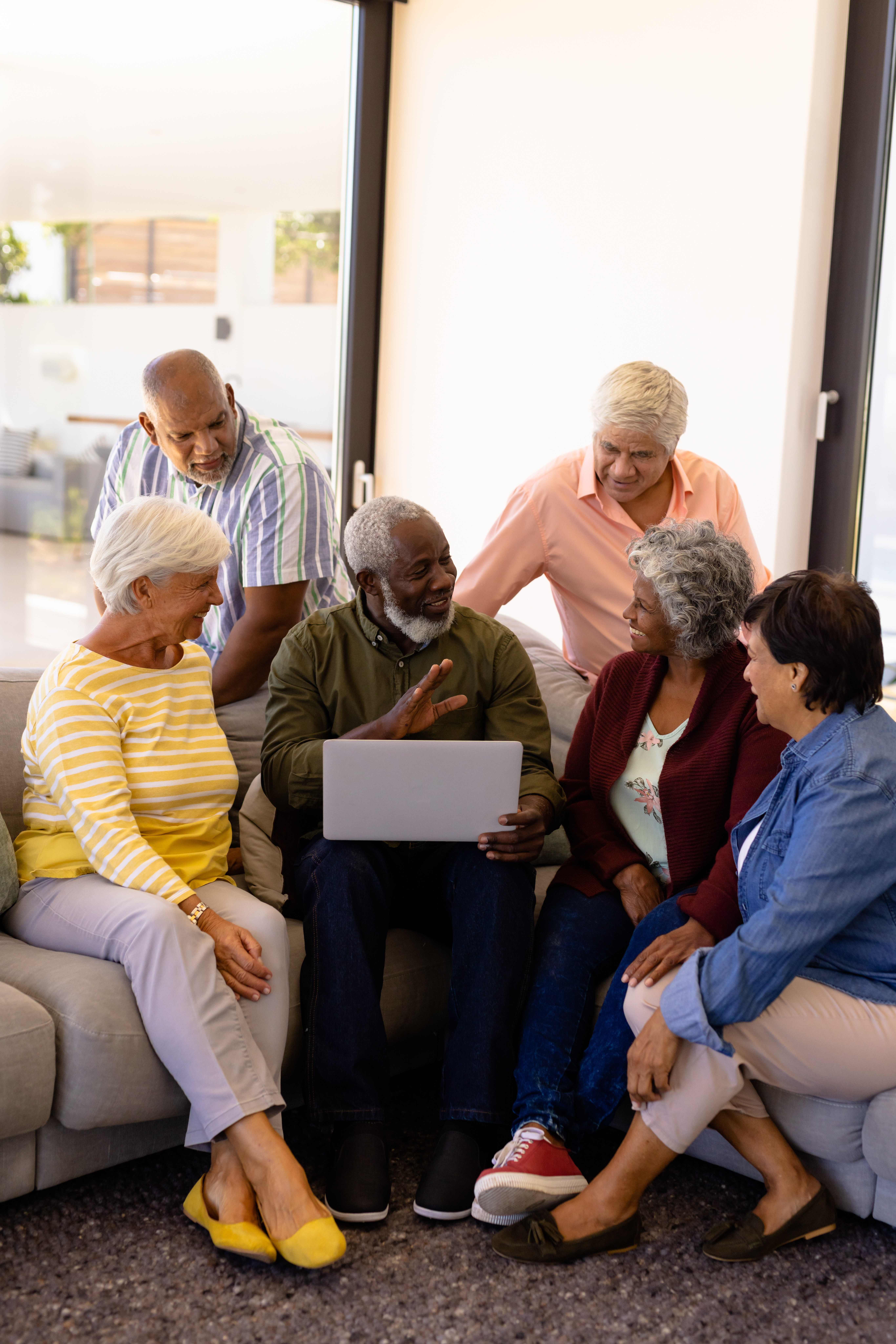 Multiracial senior man holding laptop talking with friends while sitting on sofa in nursing home